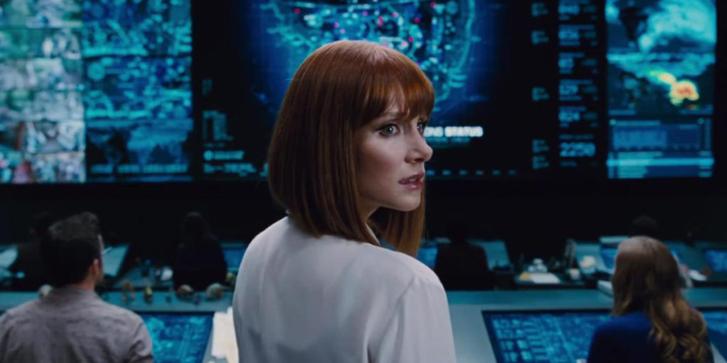 A red haired woman in white shirt in front of a large screen with a map of Isla Nublar looks over her shoulder toward the camera